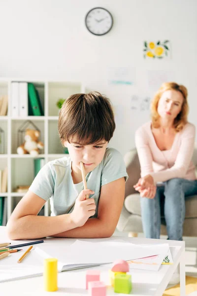 Selective focus of thoughtful kid with dyslexia holding pencil and child psychologist on background — Stock Photo