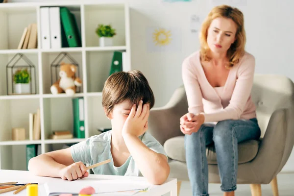 Niño triste con dislexia sosteniendo lápiz y psicólogo infantil en el fondo - foto de stock