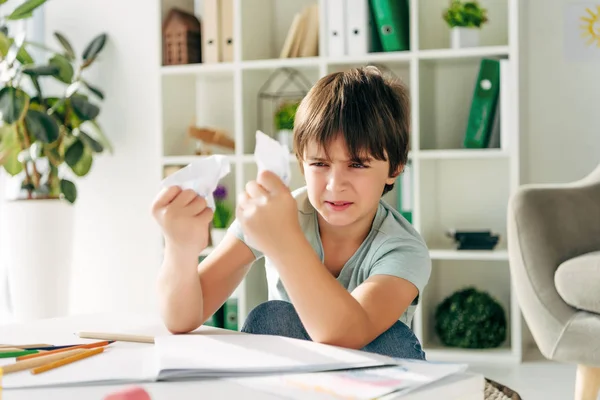 Niño irritado con dislexia sosteniendo papeles arrugados y sentado a la mesa - foto de stock
