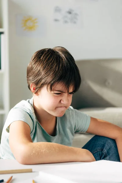 Niño irritado con dislexia sentado en la mesa y mirando hacia otro lado - foto de stock