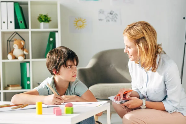 Enfant avec dyslexie tenant crayon et enfant psychologue souriant lui parlant — Photo de stock