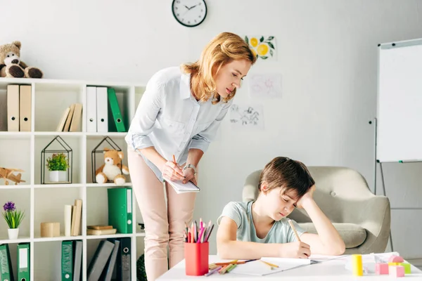 Kid with dyslexia drawing with pencil and child psychologist looking at it — Stock Photo