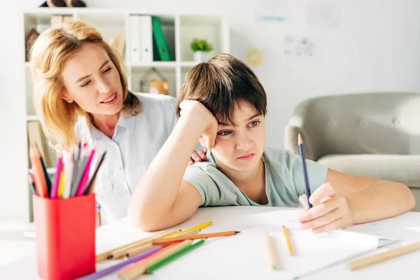 Triste niño con dislexia sosteniendo lápiz y sonriente niño psicólogo mirándolo - foto de stock