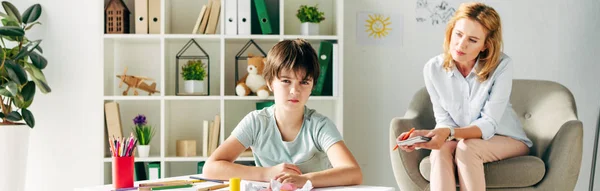 Plano panorámico de niño con dislexia mirando a la cámara y psicólogo infantil mirándolo — Stock Photo