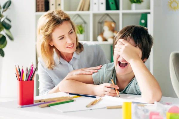 Upset kid with dyslexia obscuring face and crying, smiling child psychologist looking at him — Stock Photo
