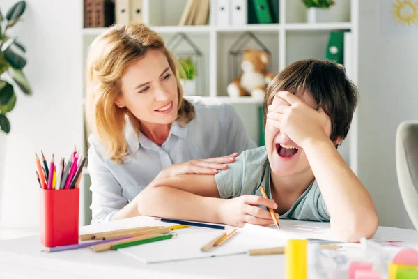 Enfant bouleversé avec dyslexie obscurcissant le visage et pleurant, enfant psychologue souriant le regardant — Photo de stock