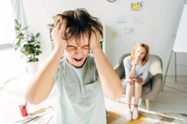Selective focus of irritated kid with dyslexia shouting and holding head — Stock Photo