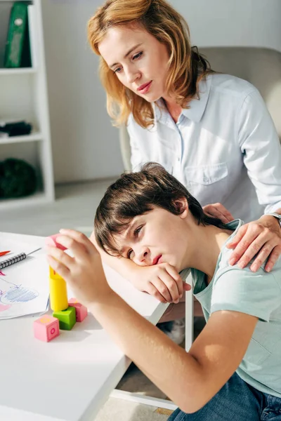 Niño con dislexia jugando con bloques de construcción y psicólogo infantil mirándolo - foto de stock