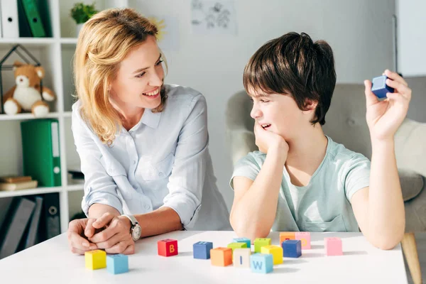 Улыбающийся детский психолог смотрит на ребенка с дислексией и сидит за столом со строительными блоками — стоковое фото