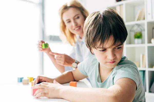 Selektiver Fokus trauriger Kinder mit Legasthenie, die wegschauen und Kinderpsychologen, die ihn anschauen und mit dem Finger auf Baustein im Hintergrund zeigen — Stockfoto