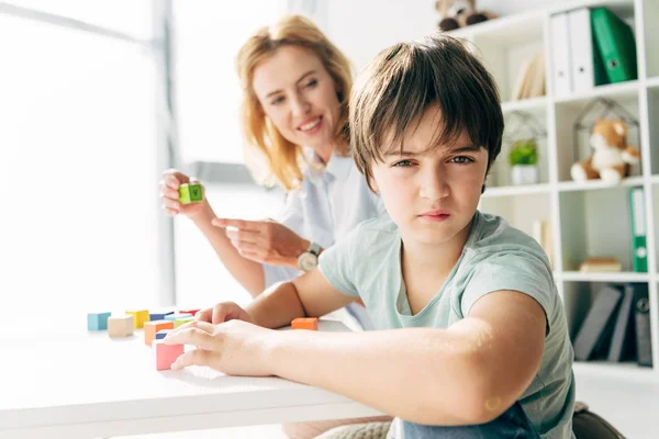 Selective focus of kid with dyslexia looking at camera and child psychologist pointing with finger at building blocks on background — Stock Photo