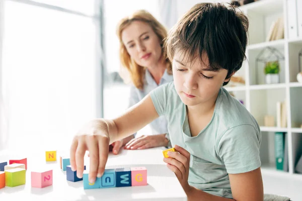 Enfoque selectivo del niño con dislexia jugando con bloques de construcción y psicólogo infantil mirándolo en el fondo - foto de stock