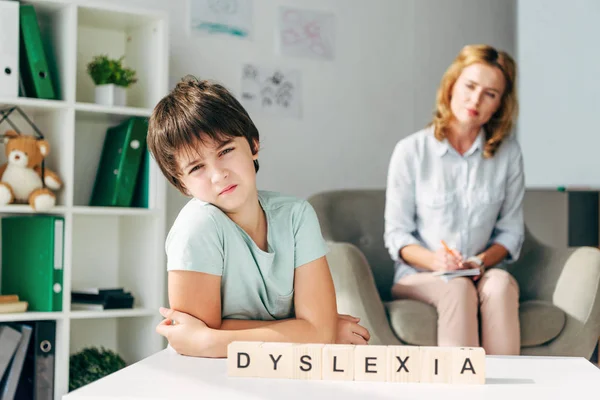 Selective focus of sad kid with dyslexia sitting at table with wooden cubes with lettering dyslexia — Stock Photo