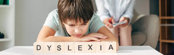 Plano panorámico de niño triste con dislexia sentado en la mesa con cubos de madera con letras dislexia - foto de stock