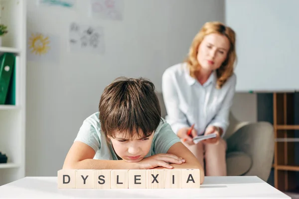 Selective focus of sad kid with dyslexia sitting at table with wooden cubes with lettering dyslexia — Stock Photo