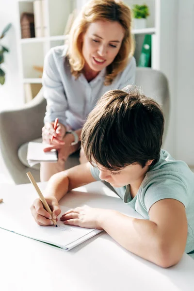 Kid with dyslexia drawing on paper with pencil and child psychologist  looking at it on background — Stock Photo