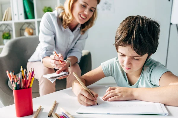 Дитина з дислексією малює на папері з олівцем та дитячим психологом, дивлячись на нього — стокове фото