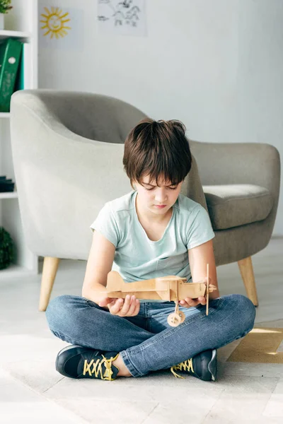 Sad kid with dyslexia sitting on floor and holding wooden plane — Stock Photo