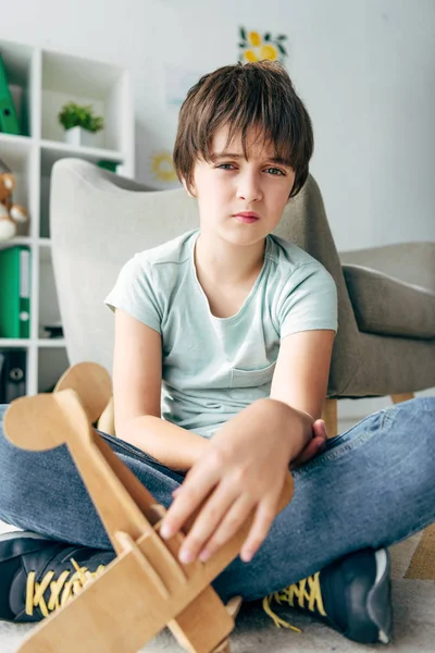 Bambino triste con dislessia seduto sul pavimento e con in mano un aereo di legno — Foto stock