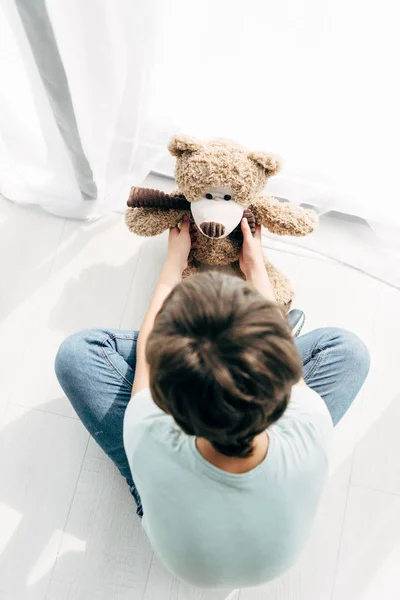 Vista superior del niño con dislexia sentado en el suelo y mirando al oso de peluche - foto de stock