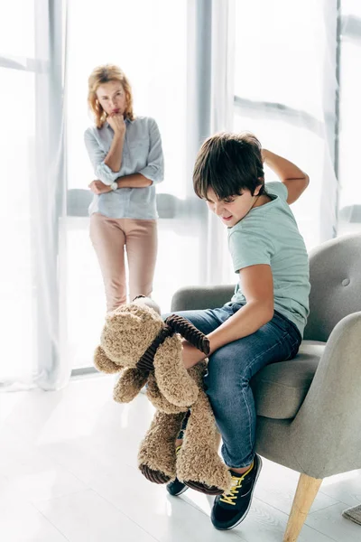 Сердитий дитина з дислексією гладить плюшевого ведмедя і дитячий психолог дивиться на нього — стокове фото