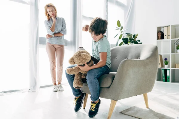 Angry kid with dyslexia punching teddy bear and child psychologist looking at it — Stock Photo