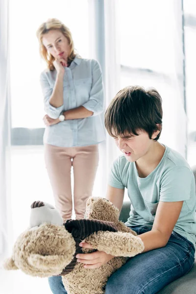 Niño triste con dislexia sosteniendo oso de peluche y psicólogo infantil mirándolo - foto de stock