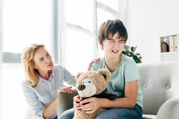 Niño triste con dislexia sosteniendo oso de peluche y psicólogo infantil hablando con él - foto de stock