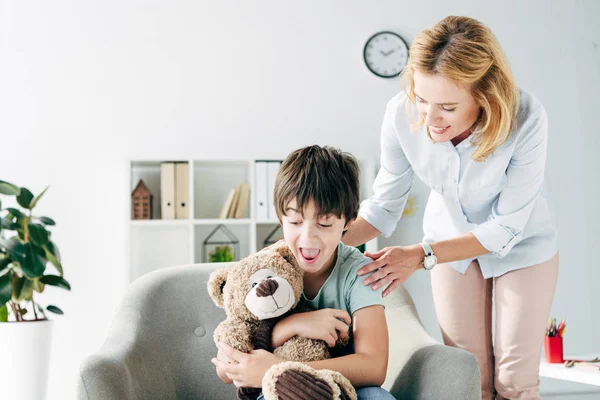 Schockiertes Kind mit Legasthenie hält Teddybär in der Hand und lächelnder Kinderpsychologe schaut ihn an — Stockfoto