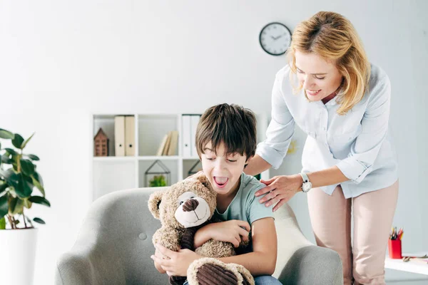 Enfant choqué avec dyslexie tenant ours en peluche et enfant psychologue souriant le regardant — Photo de stock