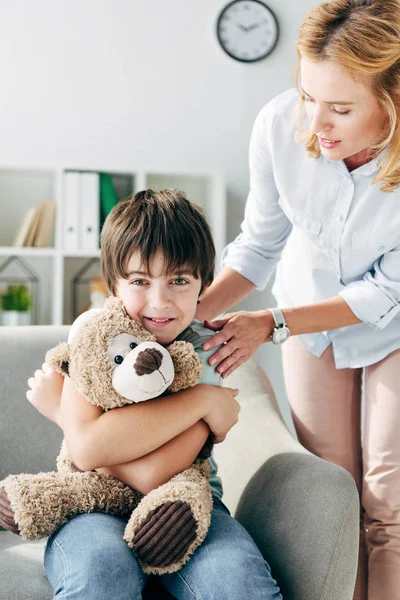 Lächelndes Kind mit Legasthenie hält Teddybär und Kinderpsychologe schaut ihn an — Stockfoto