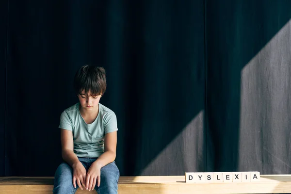 Sad kid with dyslexia sitting near wooden cubes with lettering dyslexia on black background — Stock Photo