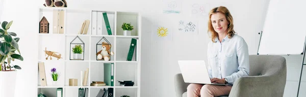 Tiro panorâmico de psicólogo infantil atraente na camisa segurando laptop e sentado em poltrona — Fotografia de Stock