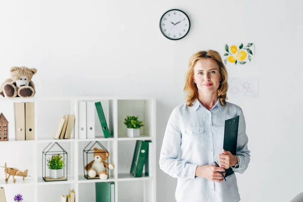 Attraktive Kinderpsychologin im Hemd blickt in die Kamera und hält Ordner in der Hand — Stockfoto