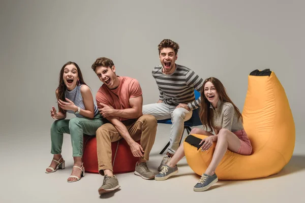 Amigos emocionais rindo e sentados em cadeiras diferentes, em cinza — Fotografia de Stock