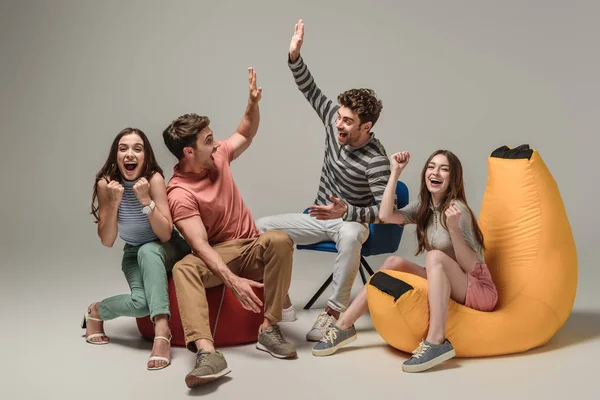 Excited friends giving highfive while sitting on different chairs, on grey — Stock Photo