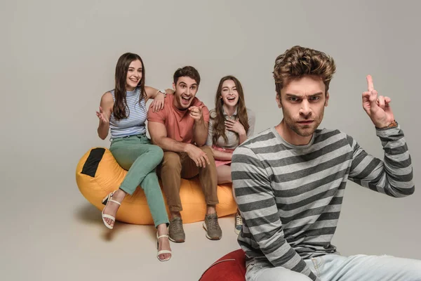 Homme en colère montrant le majeur à des amis assis sur une chaise de sac poubelle ensemble, sur gris — Photo de stock