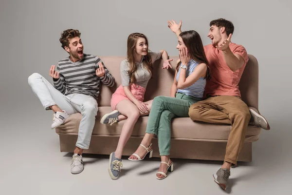 Эмоциональные молодые друзья разговаривают и сидят на диване на сером — стоковое фото