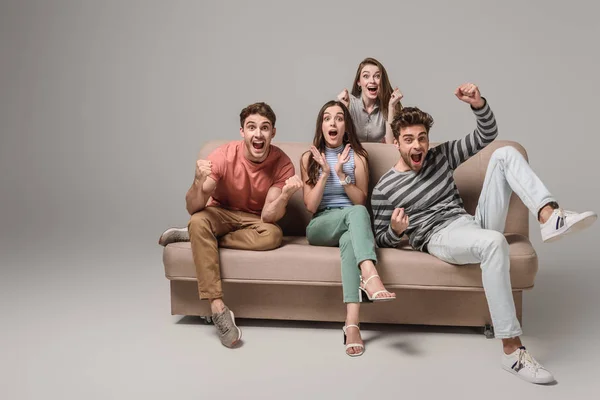 Happy young friends cheering and sitting on sofa on grey — Stock Photo