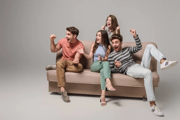 Взволнованные молодые друзья аплодируют и сидят на диване на сером — стоковое фото