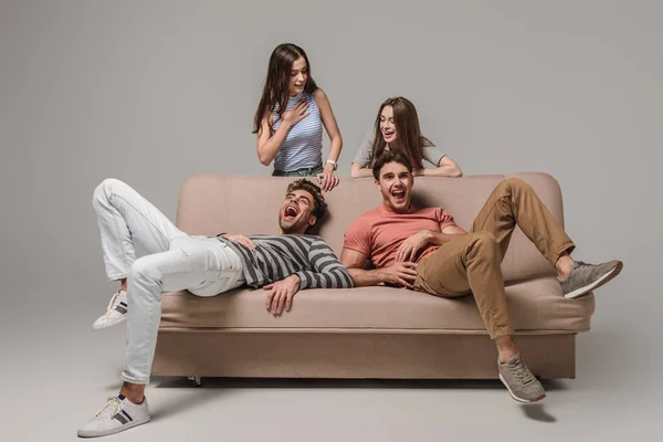 Young friends laughing and relaxing on sofa on grey — Stock Photo