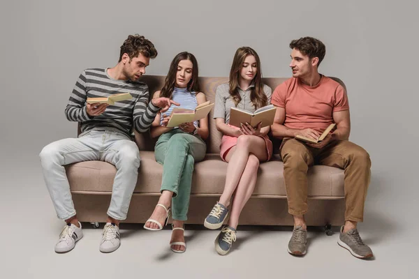 Amigos hablando y leyendo libros mientras están sentados en el sofá en gris - foto de stock