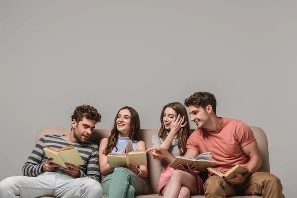 Amigos sonrientes hablando y leyendo libros mientras están sentados en el sofá en gris - foto de stock