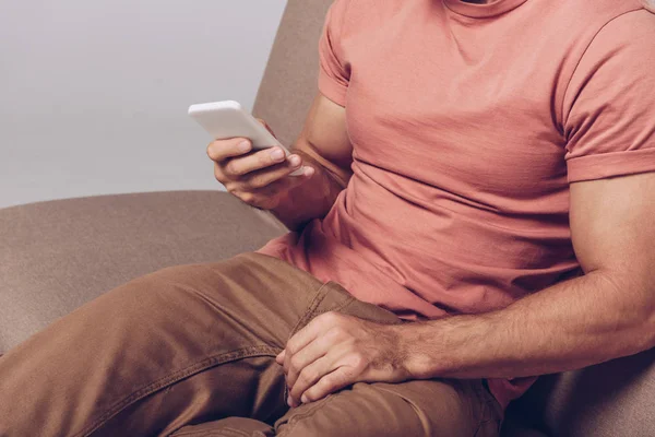 Vista parcial del hombre usando un teléfono inteligente y sentado en un sofá aislado en gris - foto de stock
