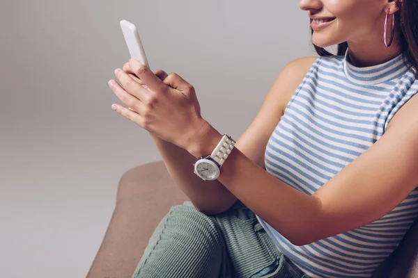 Vista recortada de la chica sonriente usando el teléfono inteligente mientras está sentado en el sofá aislado en gris - foto de stock