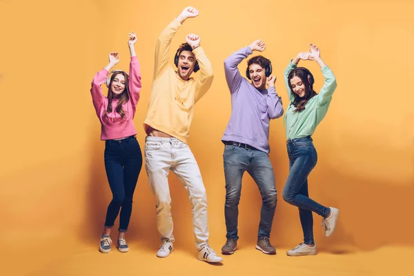 Positive friends listening music in headphones and dancing together, on yellow — Stock Photo