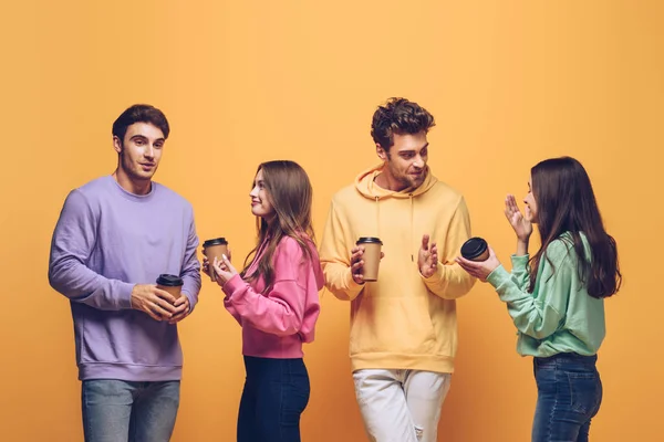 Amici felici parlando e tenendo tazze usa e getta con caffè, isolato su giallo — Foto stock