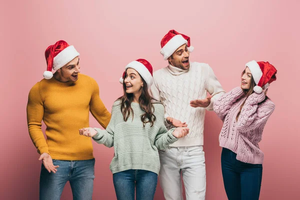 Überraschte Freunde in Pullovern und Weihnachtsmützen, die miteinander redeten, isoliert auf rosa — Stockfoto