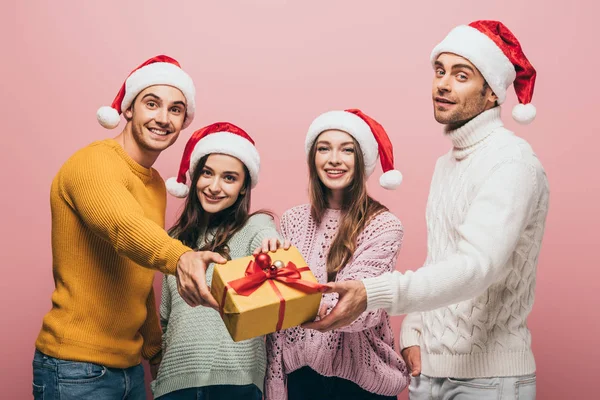 Glückliche Freunde in Weihnachtsmützen, die Ihnen eine Weihnachtsgeschenkbox schenken, isoliert auf rosa — Stockfoto