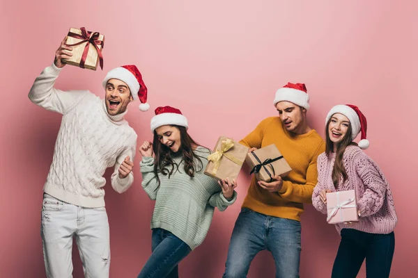 Amigos felizes em santa chapéus dançando e segurando presentes de Natal, isolado em rosa — Fotografia de Stock
